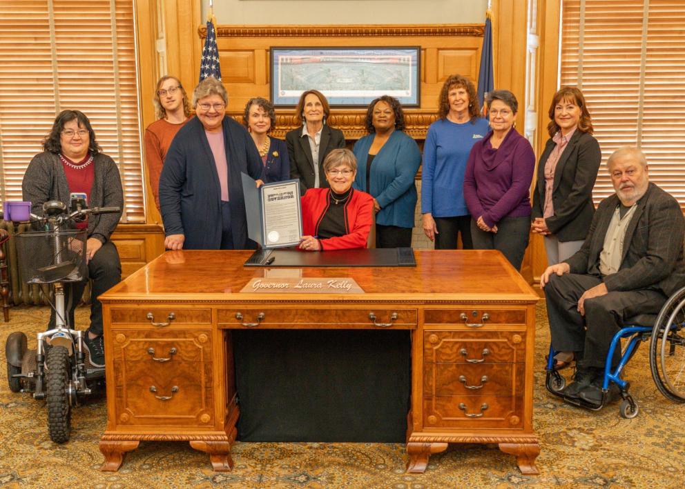A group of people stand around a wooden desk at which Gov. Kelly is seated as she holds up a signed legislative bill